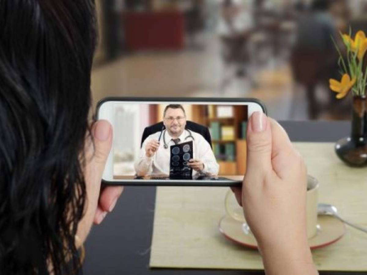 Woman sits at a cafe table and communicates with telemedicine doctor by cellphone. In touchscreen, male physician reviewing brain x-ray image. Patient has ability to reach telemedicine specialist remotely by mobile application. Horizontal indoors back side shot on cafe blurred background