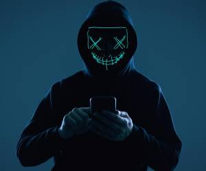 Portrait of an anonymous man in a black hoodie and neon mask hacking into a smartphone. Studio shot.