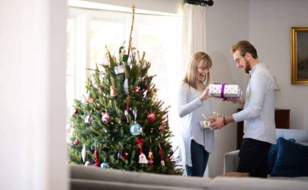 Young couple in their mid-20's and their first Christmas tree. They each hand the other a gift at the same time.