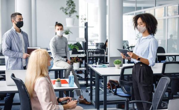 Corporate meeting and group work in modern company in office interior. African american woman manager in protective mask holding tablet, talking to workers keeping social distance during epidemic