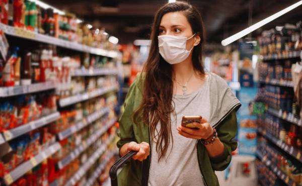 Image of a young woman in the supermarket during the coronavirus pandemic. She is checking the list of things to buy on her smartphone app.