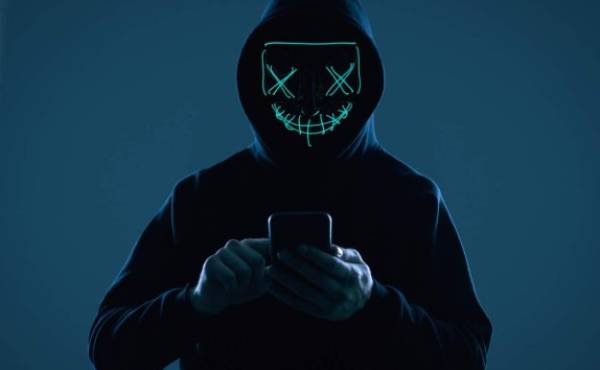 Portrait of an anonymous man in a black hoodie and neon mask hacking into a smartphone. Studio shot.